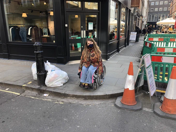 Katie, in a pink wheelchair and wearing a face mask, it at the edge of a pavement. There is no dropped kerb. Around her is lots of street clutter - bollards, signs, a-boards, bins.