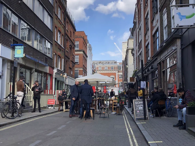 Outdoor chairs and tables on a street in soho. the pavements are blocked by a-boards and more seating.