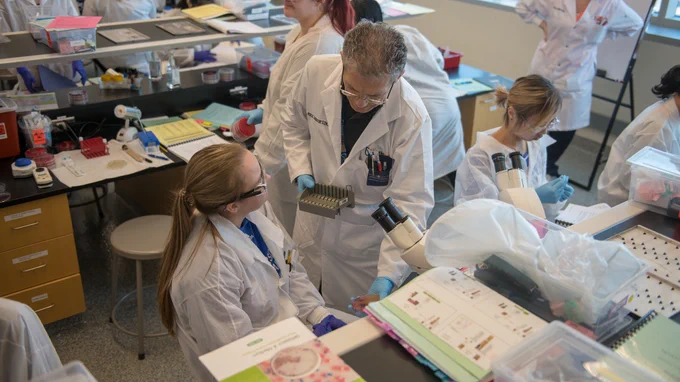 in the crowded lab, a male technician stands behind a young female with a ponytail, who is seated in front of a microscope with books to the side of it. In the background, several other students are at lab benches working on projects. 