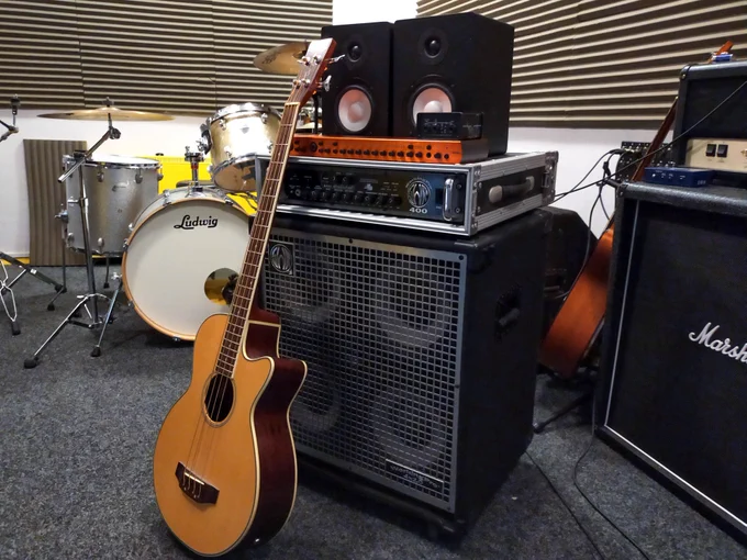 Image description: The photo shows a large bass guitar amp with four speakers and a solid metal case. On top of the amp is an orange audio interface and two studio monitor speakers. An acoustic bass guitar with a wooden finished is leant against the amp.
