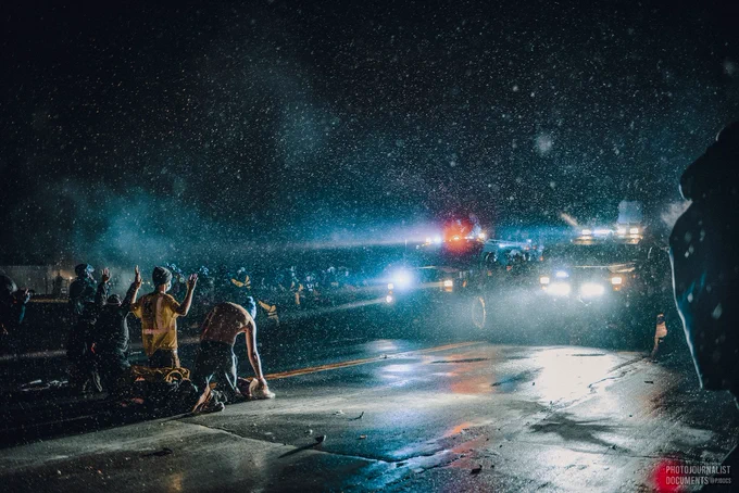 Light snow falling, four protesters kneel in the street facing two police vehicles and dozens of law enforcement officers. One protester has removed his shirt, setting it down in front of him, and two others have their hands up. (Tuesday, April 13th, 2021)
