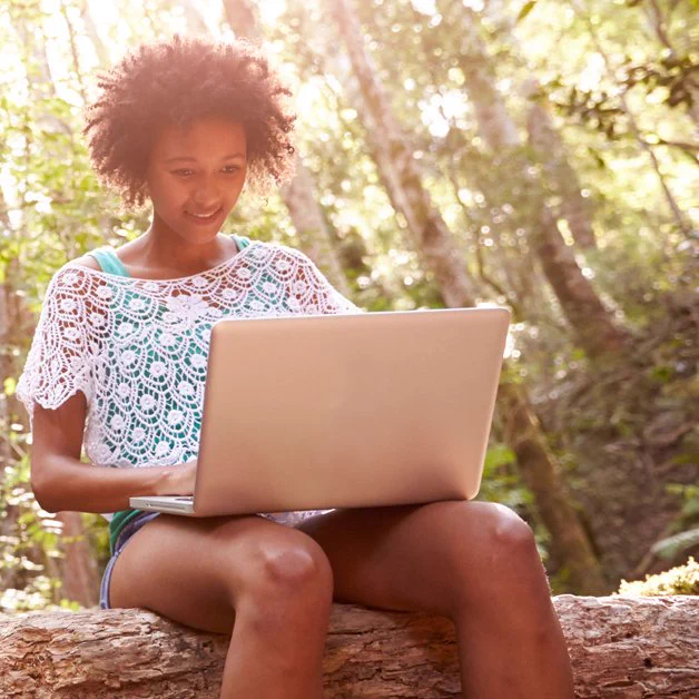 A student sits on a log in the woods with their computer on their lap