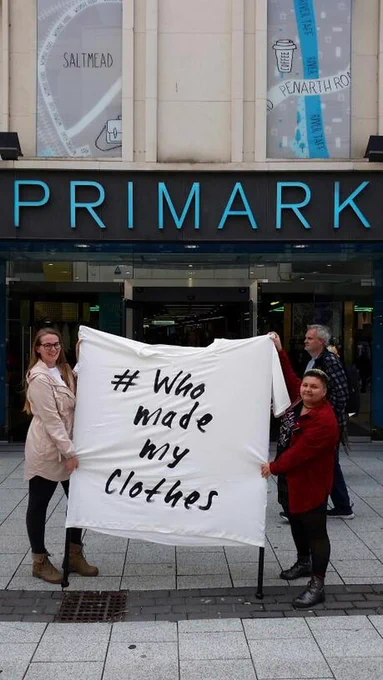 Two women are holding a large white tee that says who made my clothes outside Primark