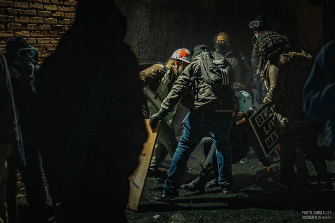 Tuesday, April 13th, 2021: Street Medics along with Black Lives Matter and Justice for Daunte Wright protesters take shelter around the corner of a nearby apartment building during a chaotic scene outside of the Brooklyn Center Police Department as they help someone wounded by police, who have used white helmets, med kits, rubber gloves, and gas masks as evidence justifying use-of-force against the crowd. Photo by independent photojournalist @pjdocs