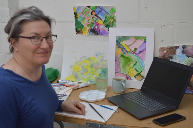 A photograph of a person sat in front of a desk. On the desk is a laptop, a cup of tea, some paints, and some paintings in watercolour.