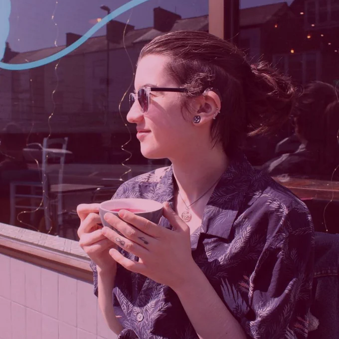 Max is sitting down outside a shop holding a drink. They are wearing glasses, hearing aids and have their hair tied back.