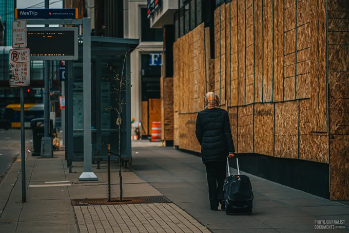 Monday, April 19th, 2021: Hastily installed plywood adorns the side of a Minneapolis building as a commuter, luggage in tow, walks to a bus stop.   Photo by independent frontline photojournalist @pjdocs
