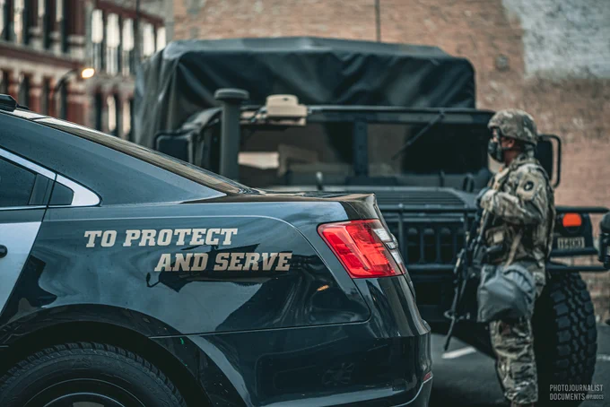 Monday, April 19th, 2021: An Anoka County Sheriff squad car, the words "TO PROTECT AND SERVE" written on the side, sits on a Minneapolis corner next to an armed Minnesota National Guard soldier and a High Mobility Multipurpose Wheeled Vehicle (Humvee). Photo by independent frontline photojournalist @pjdocs