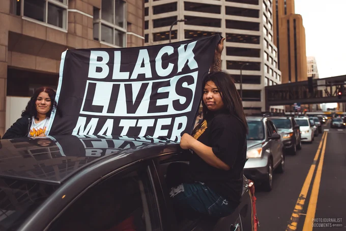 As they slowly drive down the road amidst the crowd spilling into the streets, two girls hang out the back windows of a car, holding up a flag reading "BLACK LIVES MATTER" (04/20/2021 - photo by @pjdocs) 