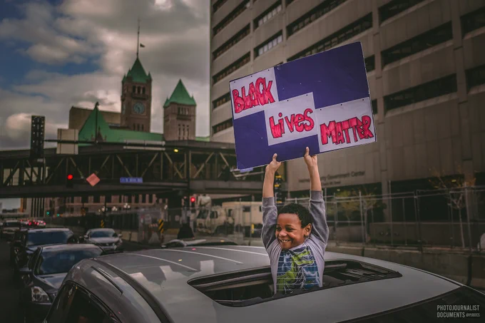A little boy stands through the opening of a sunroof, holding a sign reading "BLACK LIVES MATTER", a large smile across his face in the moments after the verdicts of Derek Chauvin came back guilty in the courthouse behind behind him (04/20/2021 - photo by @pjdocs) 