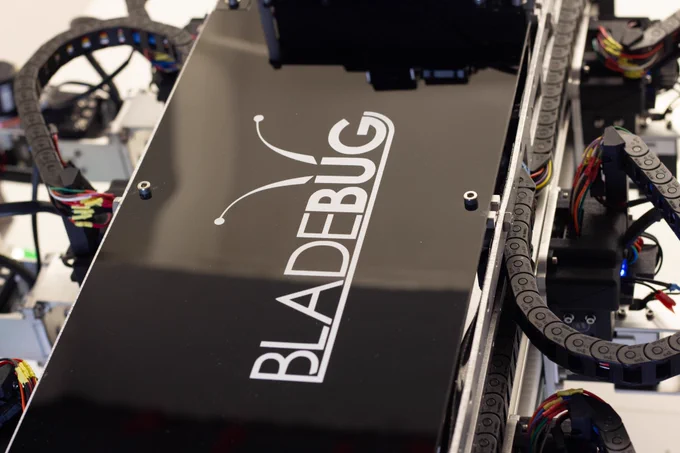 A close up of a robot, with a smooth black top and the BladeBUG robot logo vertically on the robot. There are wires and electronics on both sides.