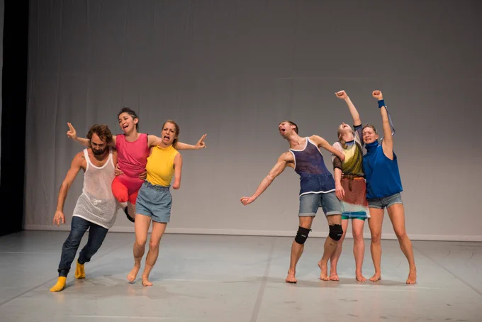 6 Candoco dancers are on a stage in groups of 3. The group on the left has 2 dancers lifting up another dancer, who has their arms wrapped around the others’ shoulders. The group on the right are huddled together, all stretching out one arm. All 6 dancers are dressed in brightly coloured clothes and have comically exaggerated expressions of pain on their faces.