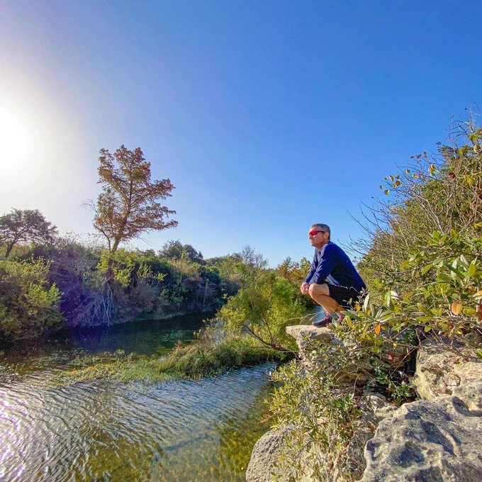 Person squatting on the edge of small cliff looking over a clear, rippling creek on a clear sunny day. 