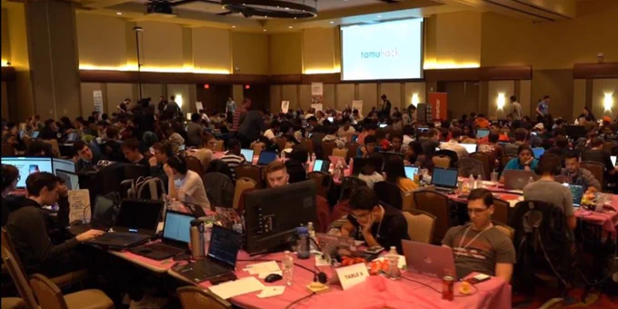 Indoors, image of a conference room with individuals sitting at long tables, dim lighting, computers out on tables, students collaborating with each other