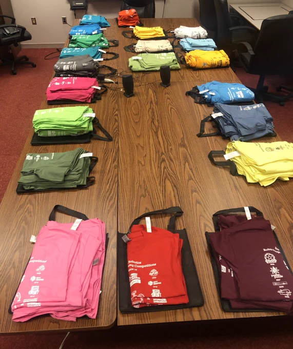 A long table with multi-coloured t-shirts organized by FIRST LEGO League team. Each team has a different colour t-shirt.