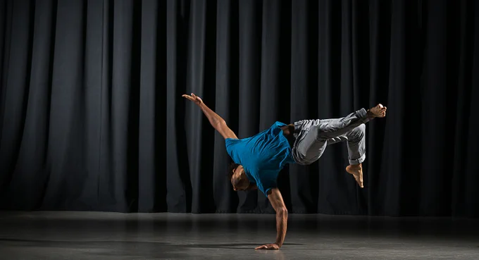 Chris is pictured mid-movement, kicking up off the floor, using his arm as support. He wears a blue t-shirt and grey jeans against a dark backdrop.