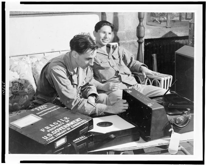 1944 black-and-white photo shows two blind men sitting and listening to a "talking book" on a record, with a Library of Congress player built for that purpose.