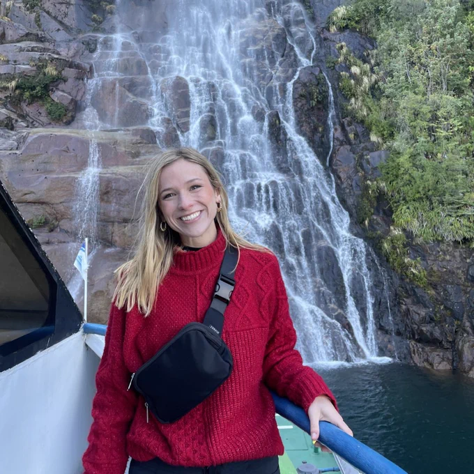 Purdue Veterinary Student Allison Jones in a red sweater standing by a boat railing, posing in front of a cascading waterfall on a rocky cliff, surrounded by greenery in Chile.