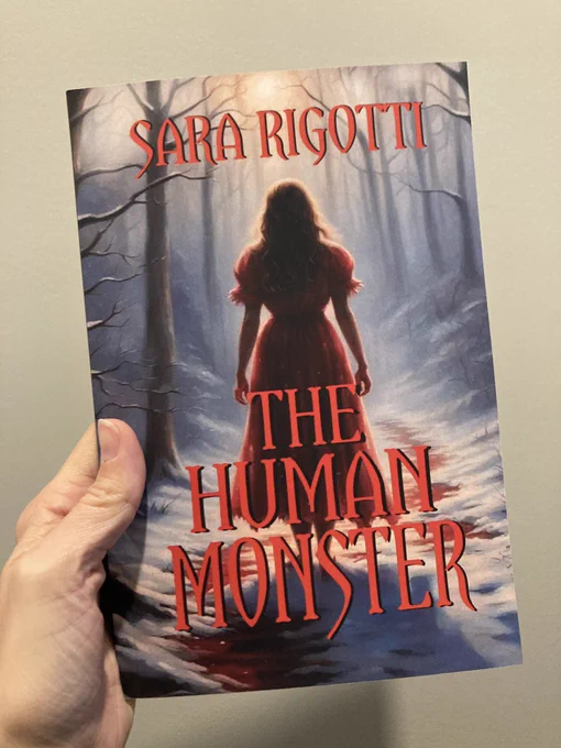 A photo of my hand holding the book, ‘The Human Monster’ by Sara Rigotti. The cover depicts an image of a woman with long wavy brown hair and wearing a red dress, walking barefoot on a snowy path in the woods, facing away from the viewer.