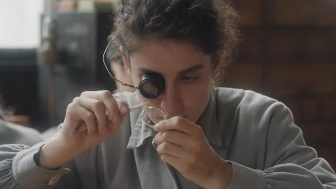 A young woman (Clara Gostynski) in a grey, long-sleeved dress shirt looks through a magnifying glass on her right eye at a watch mechanism.