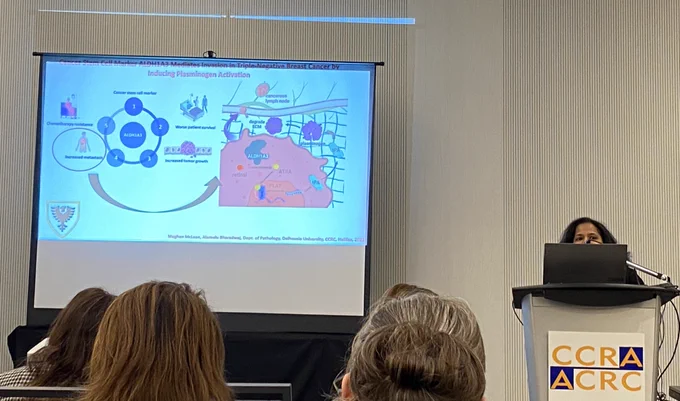 A woman peeks out from behind the podium. The slide behind her shows a diagram of cancer stem cells and metastasis.