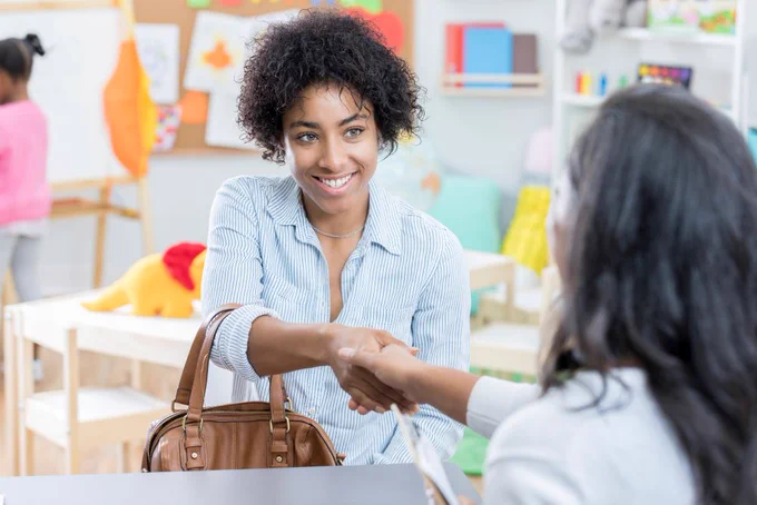 young woman smiles as she sits across a table from an unrecognizable woman in a classroom. She shakes hands