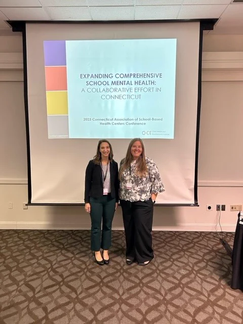CHDI's Rosie Briendel and Rebekah Behan stand together in front of the projector screen at their CASBHC presentation. They are dressed in professional clothing and smiling at the camera. The screen behind them reads, "Expanding Comprehensive School Mental Health: A Collaborative Effort in Connecticut."