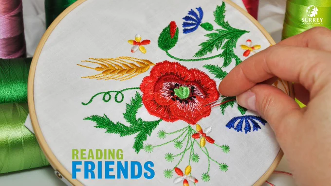 An embroidery hoop holds some white fabric, upon which someone is embroidering a floral poppy design. The blue and green Reading Friends logo and white Surrey County Council logo.