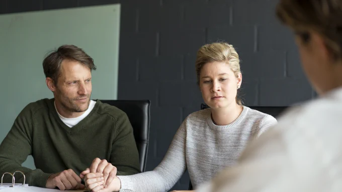 A white man with short brown hair in a side parting who is wearing a green v neck jumper is sat next to and holding the hand of a white woman with blonde hair wearing a pale round neck jumper. The man is  looking at the woman and she is looking downwards and looks thoughtful. In the foreground is the shoulder of a woman who is a healthcare professional. 