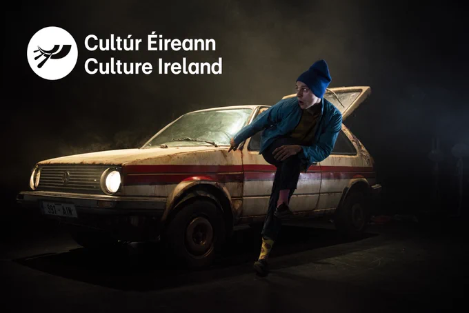 A man in blue hat jumps away from a dirty, white 1990s Volkswagen Golf car with a red strip. The car’s headlights are on and illuminate a dark space. The boot of the car is open. In the top left the white text reads ‘Cultúr Éireann Culture Ireland’, beside the Culture Ireland logo.  