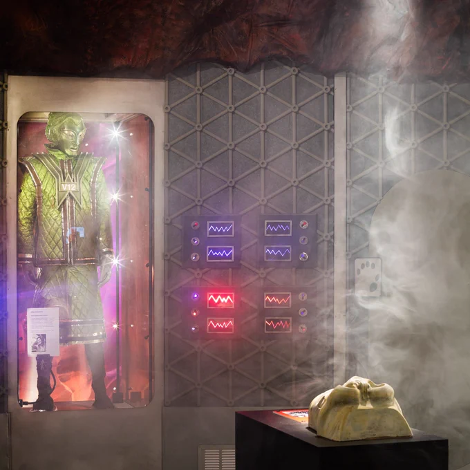 Photograph shows an exhibition space filled with dry ice, a spotlight highlighting the smoke and an object on a plinth in the foreground to the right. Behind is a display case holding a Robot of Death costume from Doctor Who, a green mask with green quilted clothes, with zigzag lights and triangular pattern on the wall to the right.