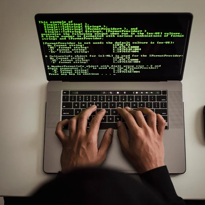 A laptop on a desk with code displayed on the screen and a set of hands typing on the laptop.