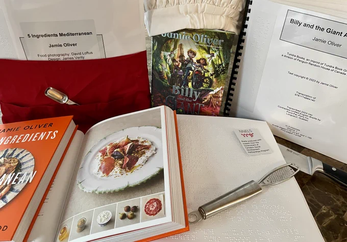 5 Ingredients Mediterranean and Billy’s Giant Adventure are arranged on a table. In the back left is a braille volume of 5 Ingredients wearing a red apron with a measuring spoon in the pocket. In the back centre is the hardcover novel wearing a chef’s hat. The braille copy of the novel is in the back right. In front, the print and braille cookbooks are open to a page featuring a fig salad. A microplane and a NNELS braille funding sticker with the red honeycomb logo rest on the braille page.