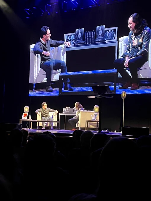 Two middle-aged men in white easy chairs on stage with a table stacked with books between them. There is a screen with the stage scene repeated above them, but much larger. 