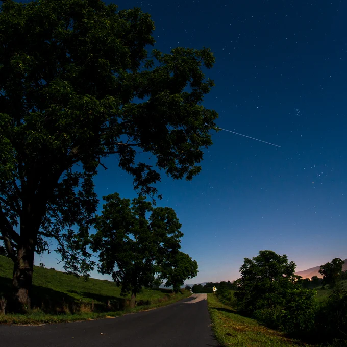 A country road at dawn, flanked by small trees and a gently-sloping hill. Above a pinkish horizon, a dark blue sky dominates the right side of the image, with a thin white line, representing a long exposure of the International Space Station, standing out amidst gently sparkling stars. Photo credit: NASA/Bill Ingalls