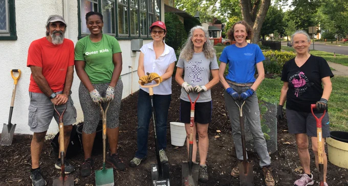 Six smiling people stand outside next to a house. They are all wearing garden gloves and holding shovels.