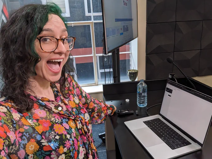I am standing on a stage, ready to do a talk. You can see my laptop in presentation mode on a dark desk with a window behind it. 
I'm wearing a floral dress and grinning at the camera with my mouth open