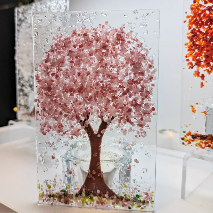 Fused glass pink cherry blossom tree candle shade on a shelf with orange Autumn & white winter trees in the background. 