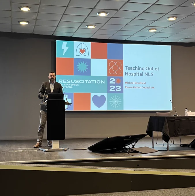 Man speaking at a podium in front of a screen reading “Teaching Out of Hospital NLS, Michael Bradfield, Resuscitation Council UK”