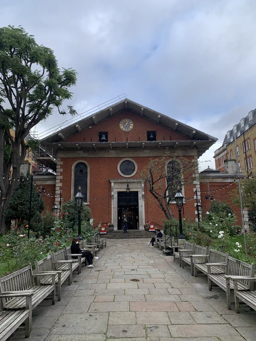 This is a medium distance photo of the exterior of a red-brick church without a spire. There are benches lining a pathway which leads to the front door of the church.