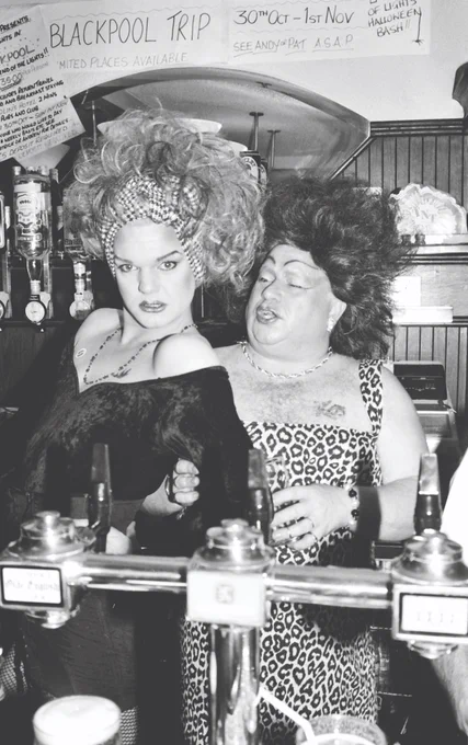A black and white photo of two drag queens posing for the camera behind a pub bar.
