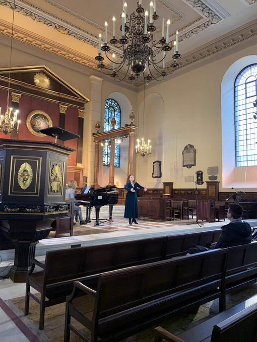 This is a photo of Joanne, singing inside St Paul’s Church Covent Garden. There are church pews in the foreground and a raised pulpit to the left.