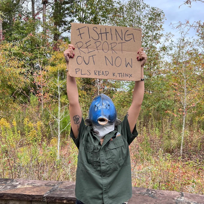 WRD employee wearing a fish mask while holding up a sign that reads 'FISHING REPORT OUT NOW. PLS READ. K, THNX.'