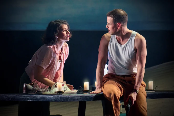 A woman in a pink top leans on a candlelit table outdoors, arguing with a man in a white top who holds a gun.