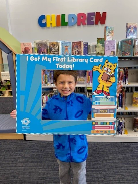 Thomas from Mountain Road Library holding up a "I got my first library card today!" frame, standing in front of bookshelves in the children's section of the library.