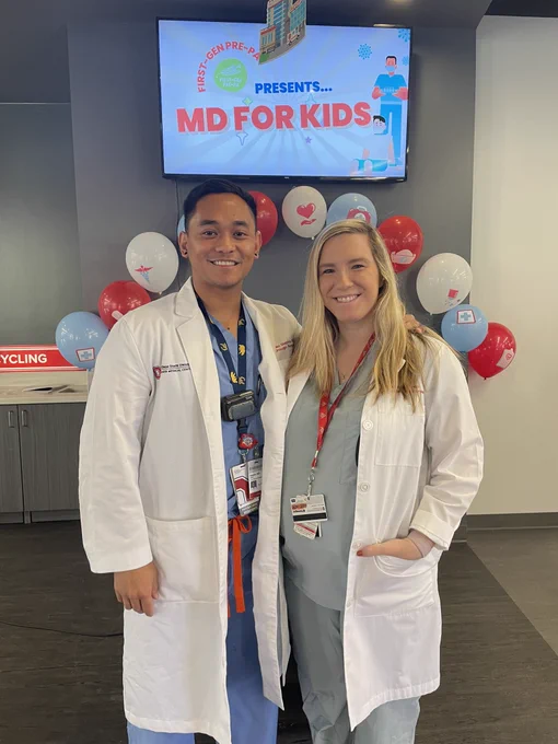 Resident Allen Siapno and Faculty Tasha Posid wear scrubs and white coats in front of an MD For Kids sign.