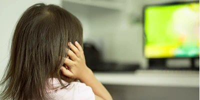 Young girl with long dark hair sits with back to camera, right hand cupped over ear, watching bright lime green computer screen that is out of focus and blurry in background.