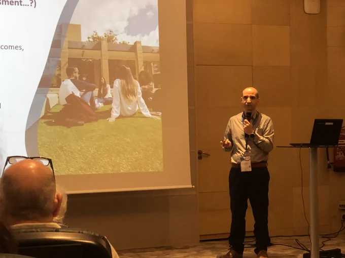 Image of Dario Pirotta standing holding microphone. Screen visible
