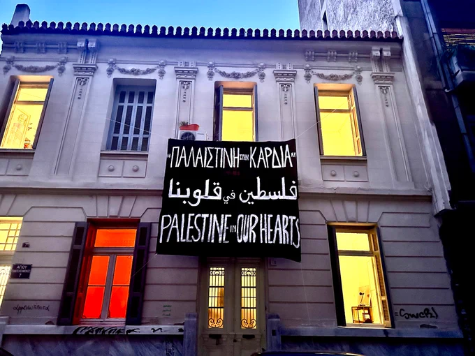A banner reading “Palestine in our hearts” (in Greek, Arabic, and English) hangs outside the feminist Autonomous Centre for research in Athens at dusk 