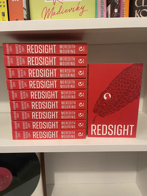 Advance reader copies of REDSIGHT by Meredith Mooring in a stack on a white bookshelf. One copy is face-out showing the bright red cover with an image of a snake outlined in a slightly darker shade.
